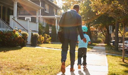 Father walks his son to school along a quiet suburban street filled with green lawns and trees.