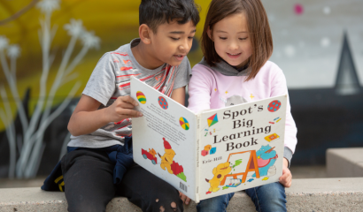 A elementary school aged boy and girl sit outside, staring at a large book.