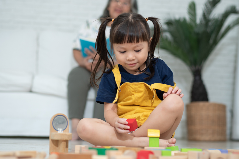 A little toddler girl, who is surrounded by wooden, colorful blocks, picks up a red block, staring at it.