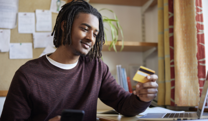 A male college student holds a credit card and his cellphone as he sits at his computer desk.