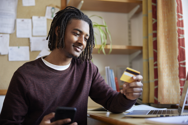 A male college student holds a credit card and his cellphone as he sits at his computer desk.