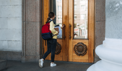 A mixed-race college students stands outside the doors of a college building. She attempts to open the double doors.