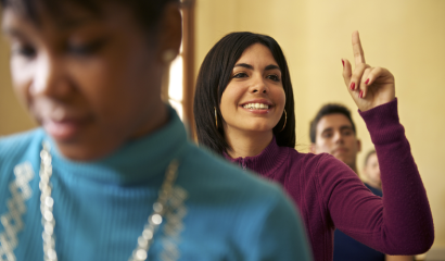 A female student in the middle of a row of desks raises her hand.