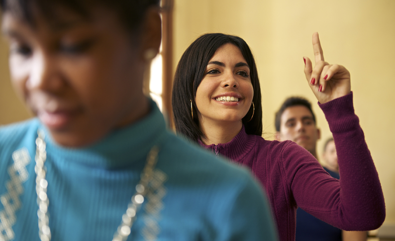 A female student in the middle of a row of desks raises her hand.