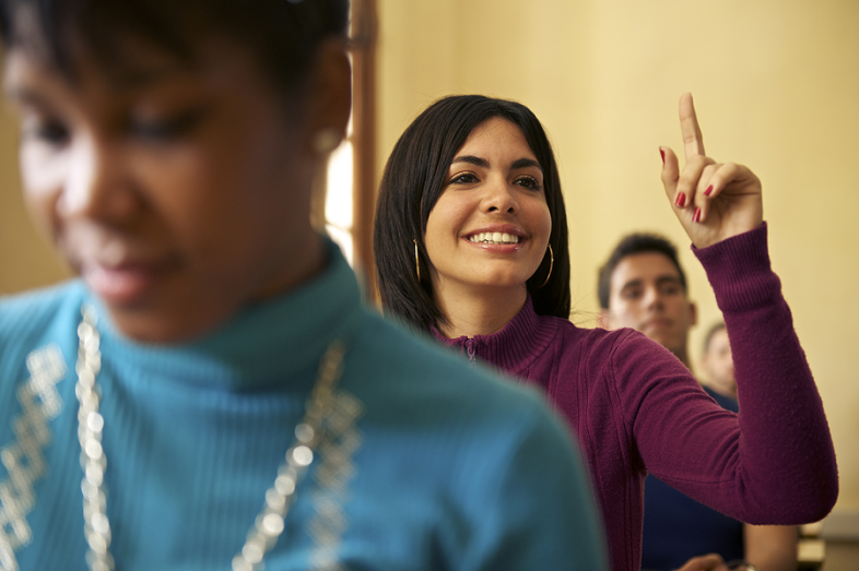 A female student in the middle of a row of desks raises her hand.