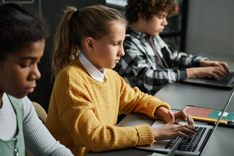 Three children of different ethnicities sit at a long desk and focus doing their schoolwork on laptop computers.