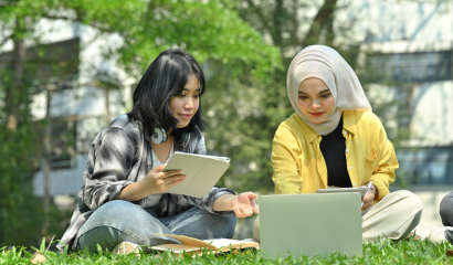 Two Asian college students sit outdoors on grass, working on an assignment