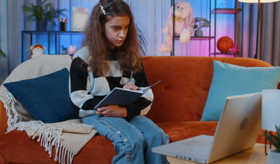 A preteen girl sits in her colorful bedroom studying and looking at her laptop screen.