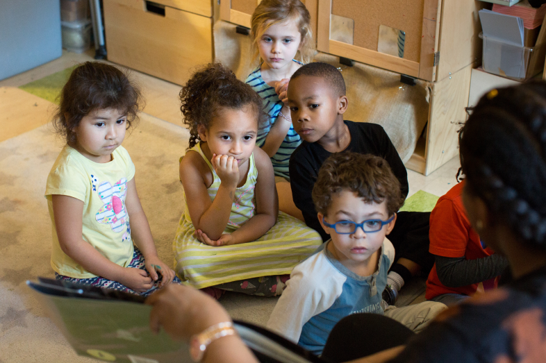 Children sit on the floor, listening to their teacher read.