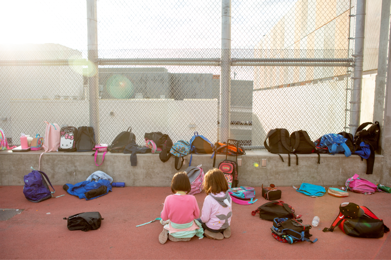 Two small elementary-aged children are outside their school, surrounded by the backpacks of their classmates.