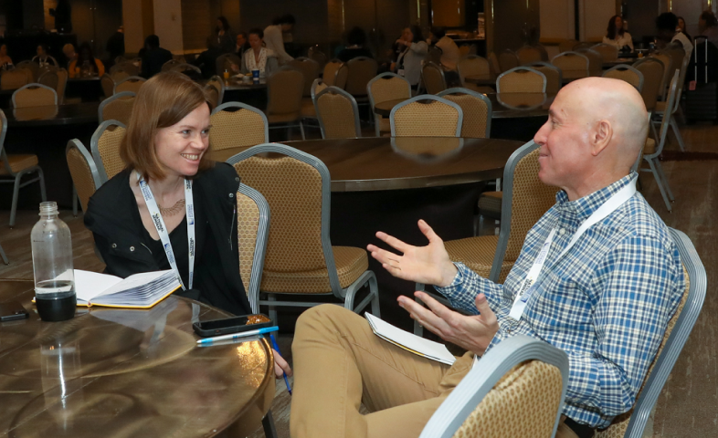 A white woman and white man speak while sitting at a table.