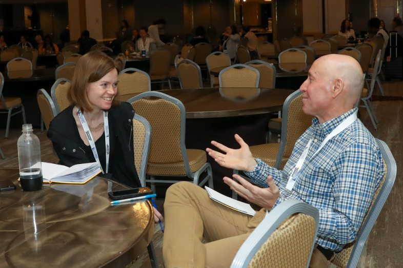 A white woman and white man speak while sitting at a table.