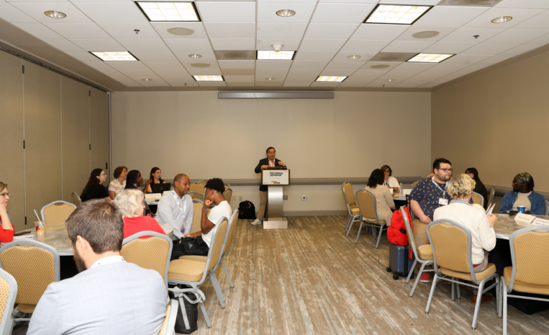 A male stands at a podium in front of a room filled with people sitting at roundtables.