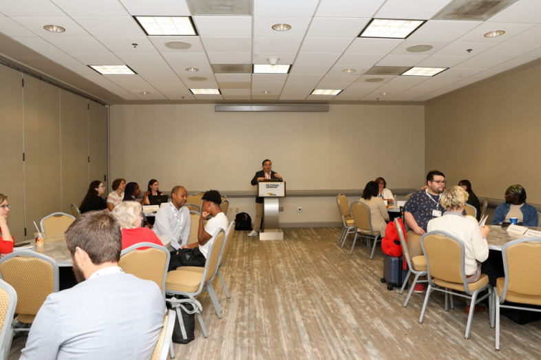 A male stands at a podium in front of a room filled with people sitting at roundtables.