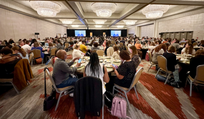 Groups of people sit at roundtables in a meeting hall, eating meals.