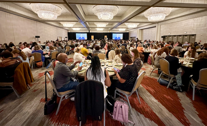 Groups of people sit at roundtables in a meeting hall, eating meals.