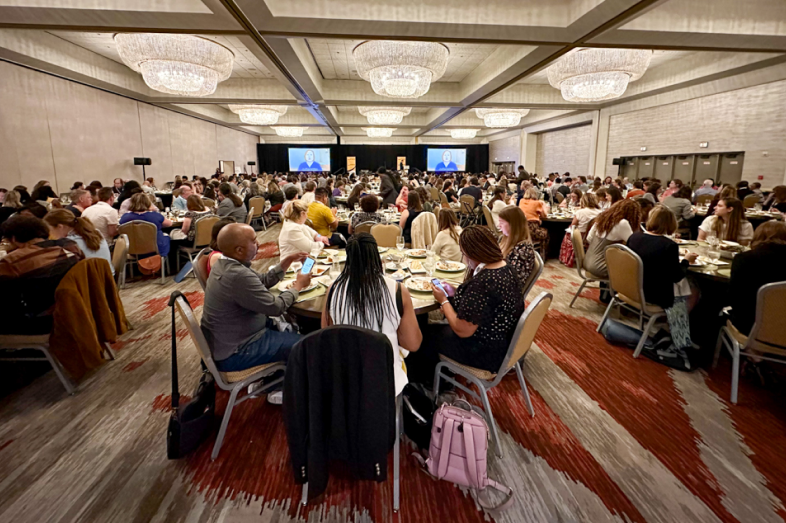 Groups of people sit at roundtables in a meeting hall, eating meals.