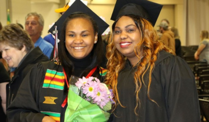 Two Black female college graduates pose side by side and smile for a photo.