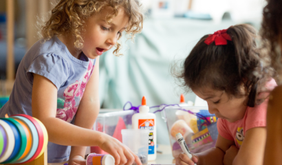 Two preschoolers color and look at artwork on a desk.