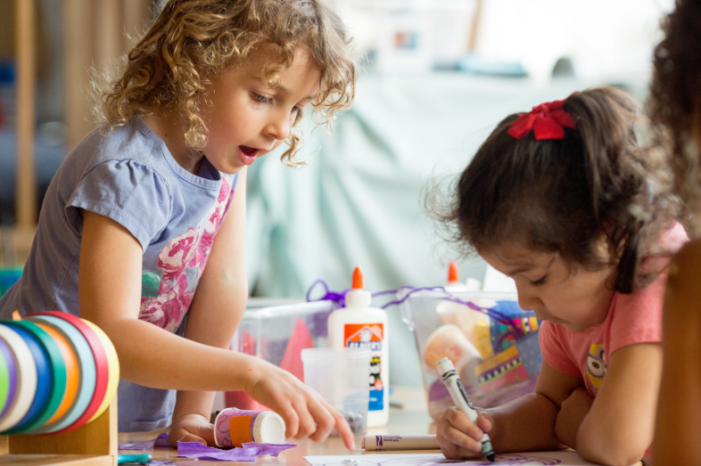 Two preschoolers color and look at artwork on a desk.