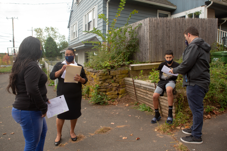 Three adults stand outside talking. A preteen sits outside his home.