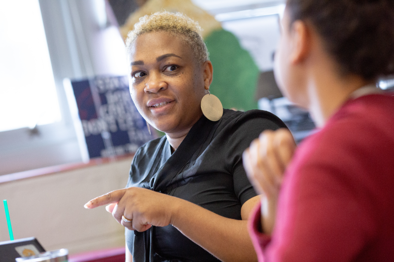 A woman points as she talks to another woman sitting next to her.